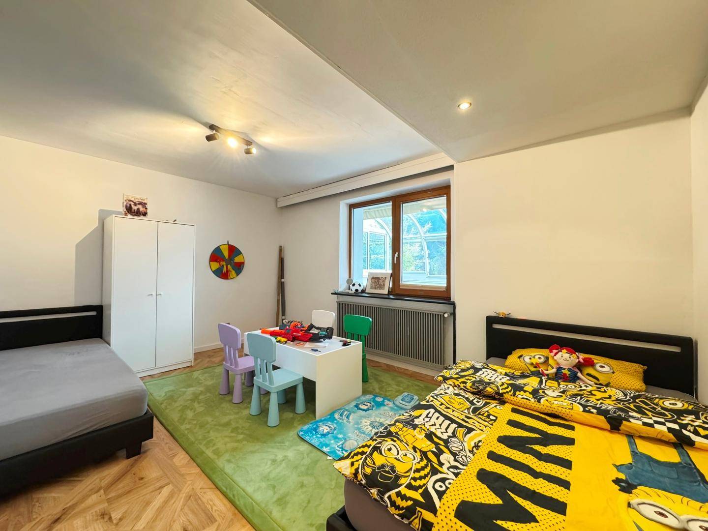 Kinderzimmer mit zwei Betten, Spieltisch und einem Fenster mit Blick ins Grüne.