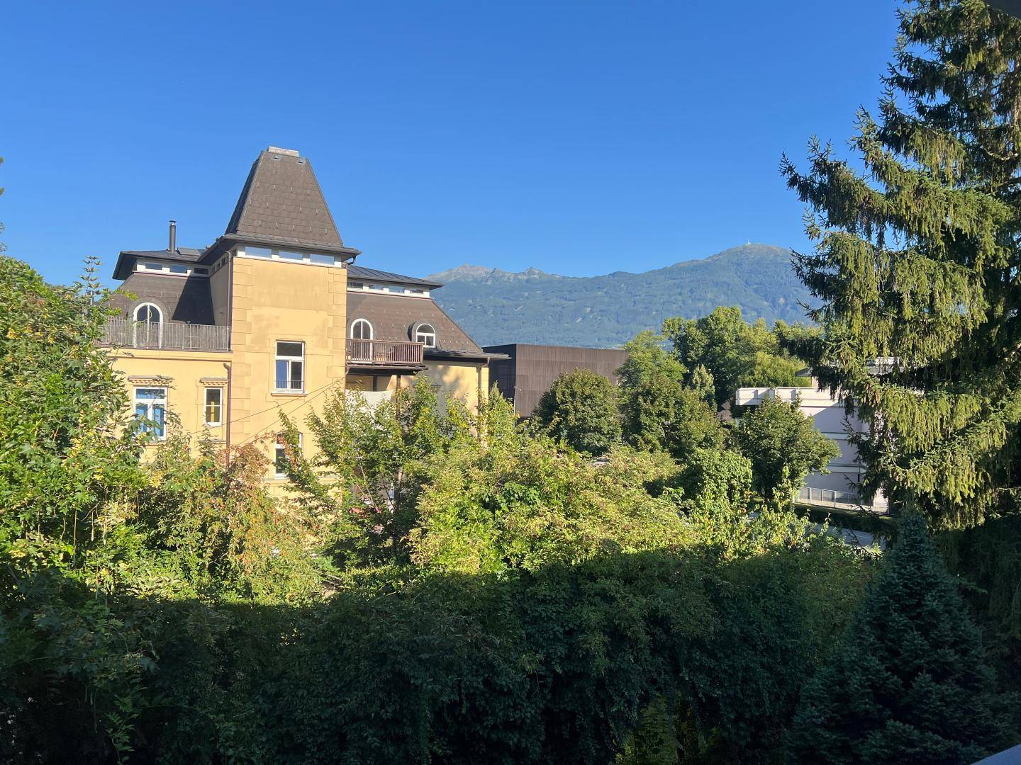 Historisches Gebäude mit markantem Dach, umgeben von Bäumen und Bergblick im Hintergrund.