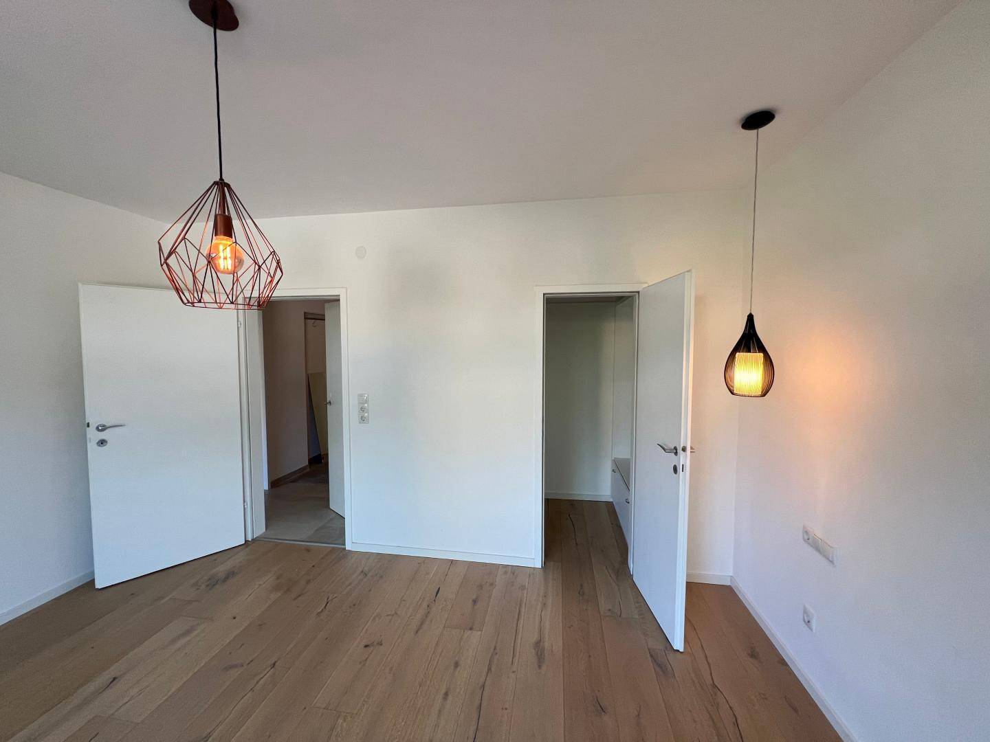 Leerer Raum mit Holzboden, weißen Wänden und modernen Hängelampen, bereit zur Einrichtung.