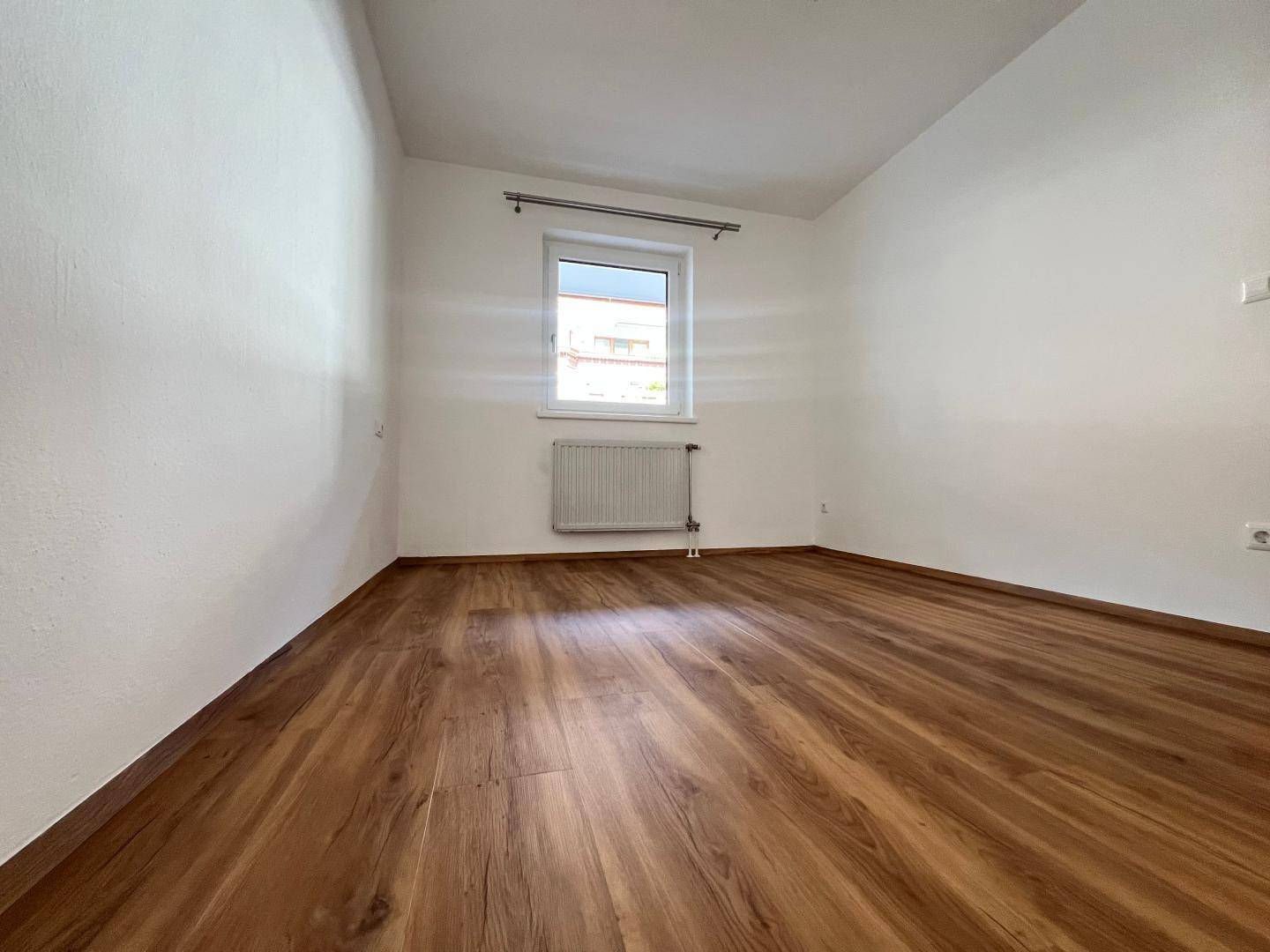 Helles Zimmer mit Laminatboden und einem Fenster, das viel Tageslicht hereinlässt.