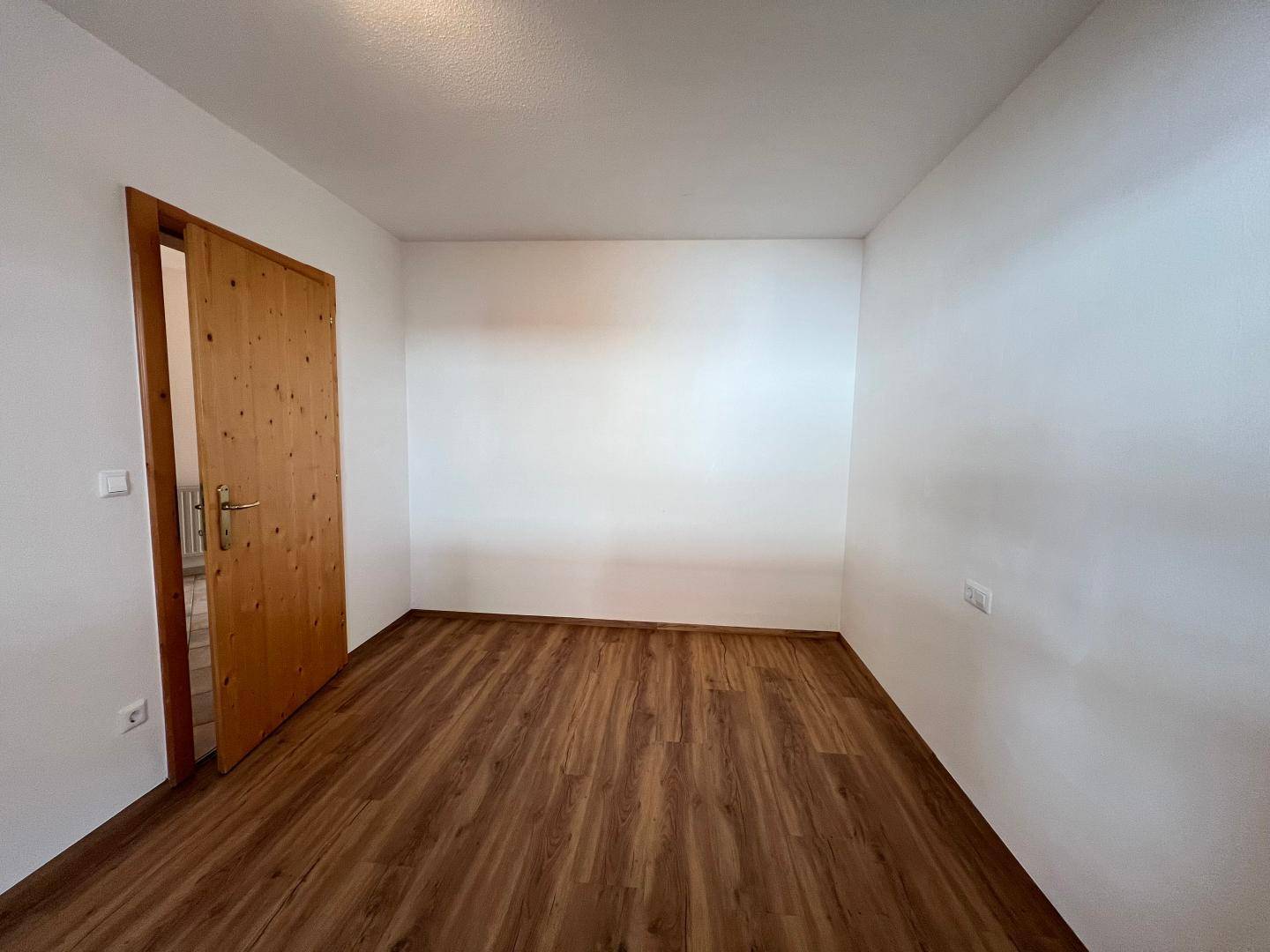 Leeres Zimmer mit Laminatboden und schlichter weißer Wandgestaltung, bereit zur Einrichtung.