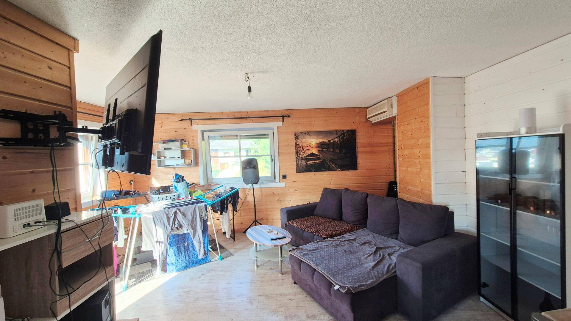 Gemütliches Wohnzimmer mit Holzwänden, Sofa und großem Fernseher, hell durch Fenster.