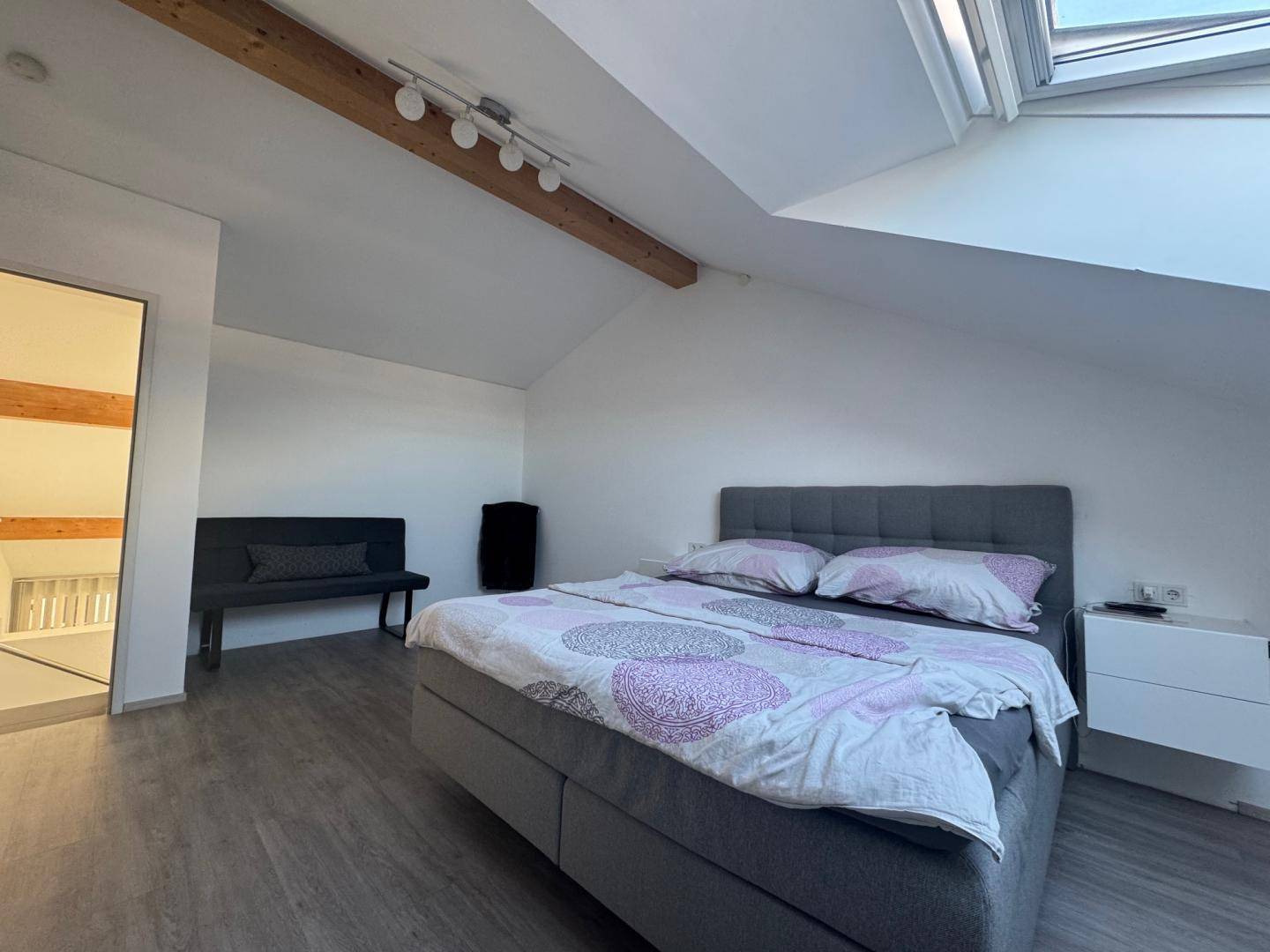 Helles Schlafzimmer mit Dachfenster, Holzbalken und Blick auf den angrenzenden Flur.