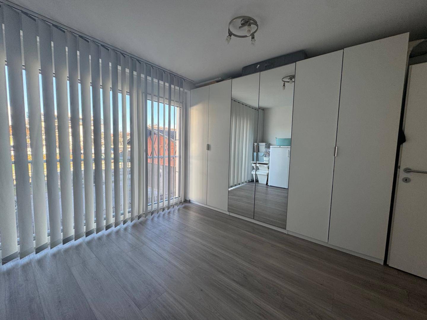 Helles Zimmer mit großen Fenstern, vertikalen Jalousien und integrierten Spiegelschränken.