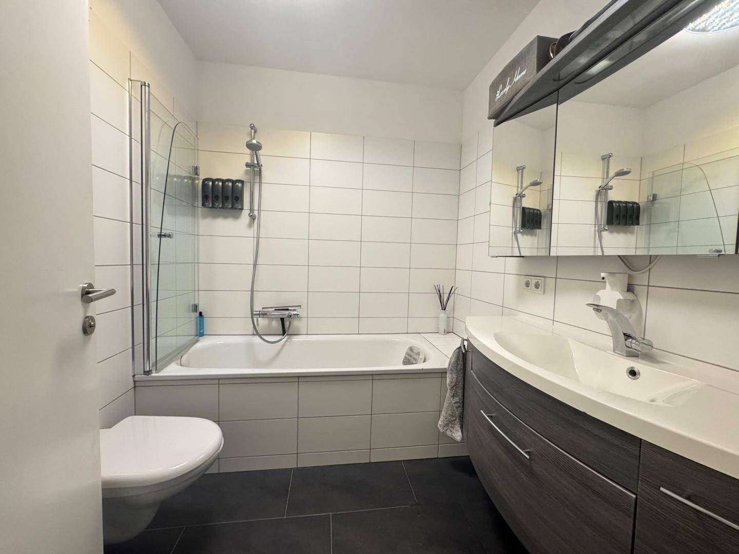 Modernes Badezimmer mit Badewanne, Duschwand, wandhängendem WC und Waschtisch mit Spiegelschrank.