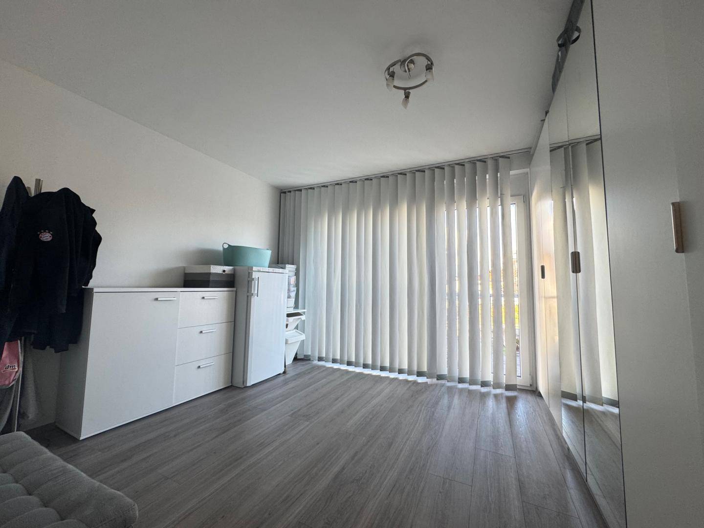 Geräumiges Zimmer mit viel Tageslicht, vertikalen Jalousien und praktischen Einbauschränken.