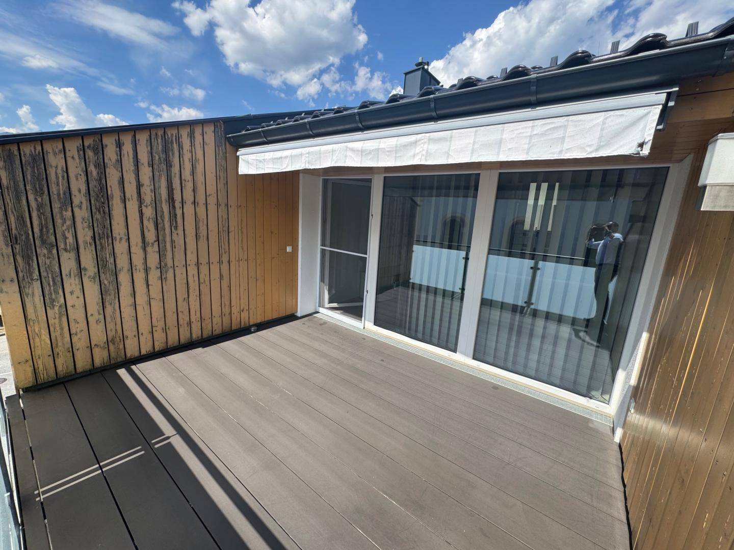 Heller Balkon mit Holzboden, Glasgeländer und großer Schiebetür, ausgestattet mit einer Markise.