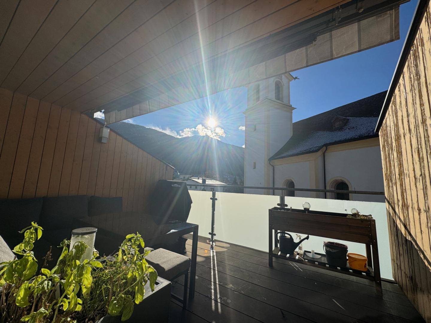 Sonniger Balkon mit Sitzgelegenheiten, Pflanzen und herrlichem Blick auf die Berge und die Kirche.