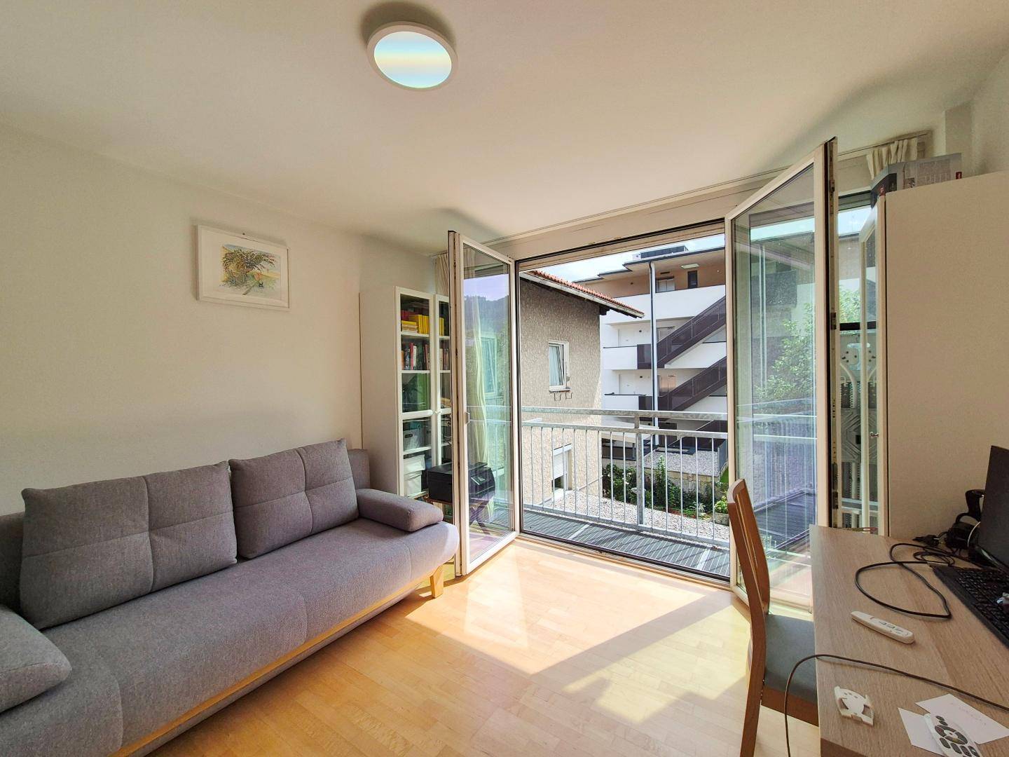 Heller Raum mit grauem Sofa, Schreibtisch und großen Glastüren, die zu einem kleinen Balkon führen.