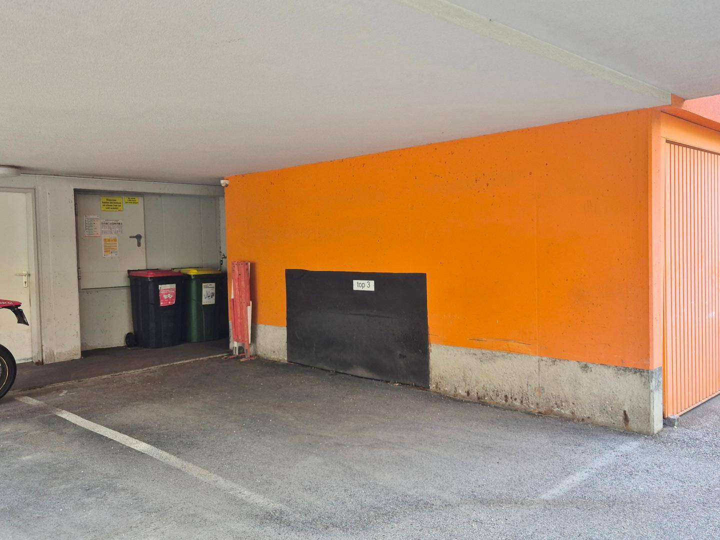 Überdachter Parkbereich mit orangefarbener Wand und Zugang zu einer Garage sowie Mülltonnen.