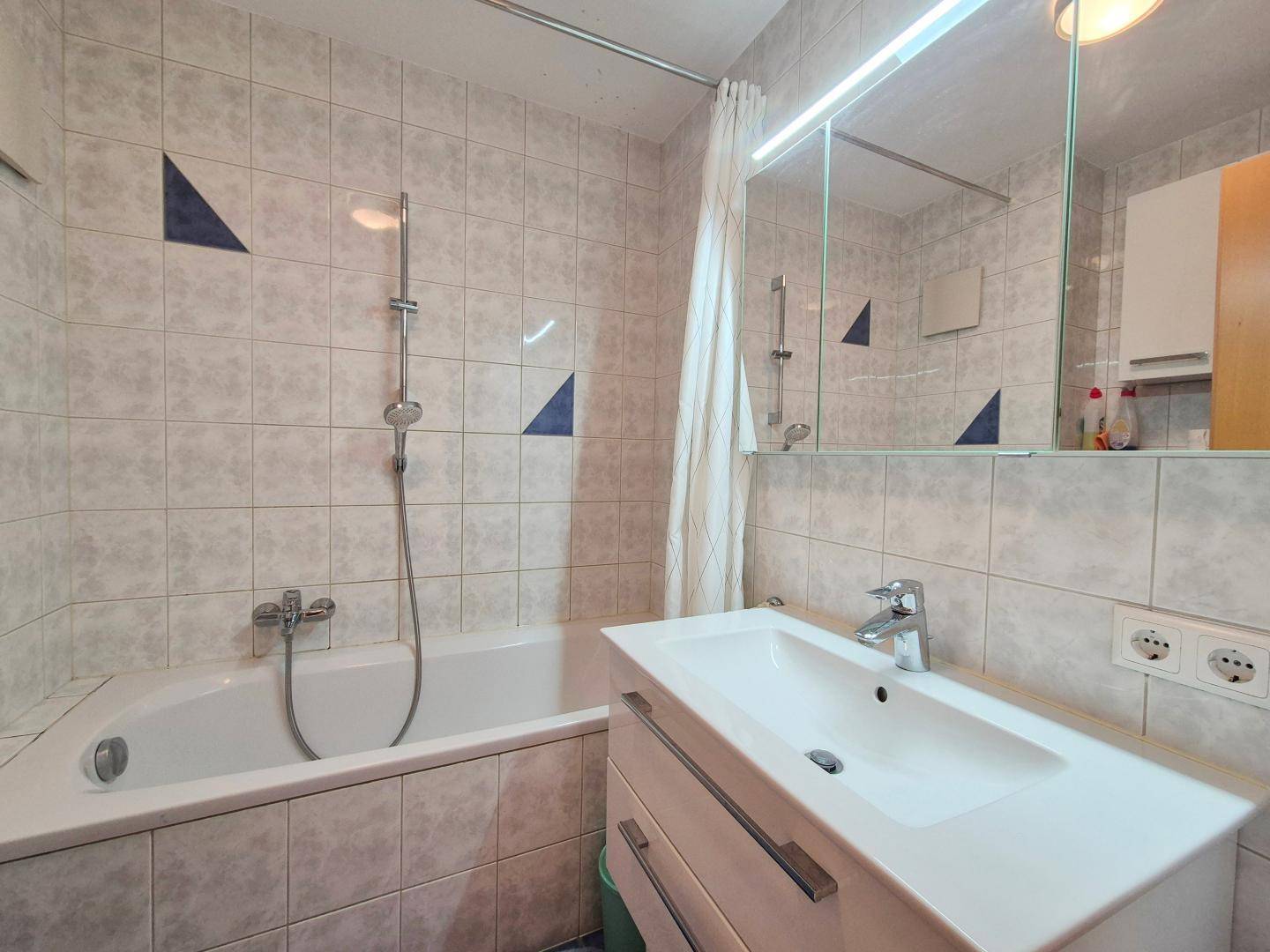 Badezimmer mit hellgrauen Fliesen, Badewanne mit Duschfunktion und einem Waschbecken mit Spiegelschrank.