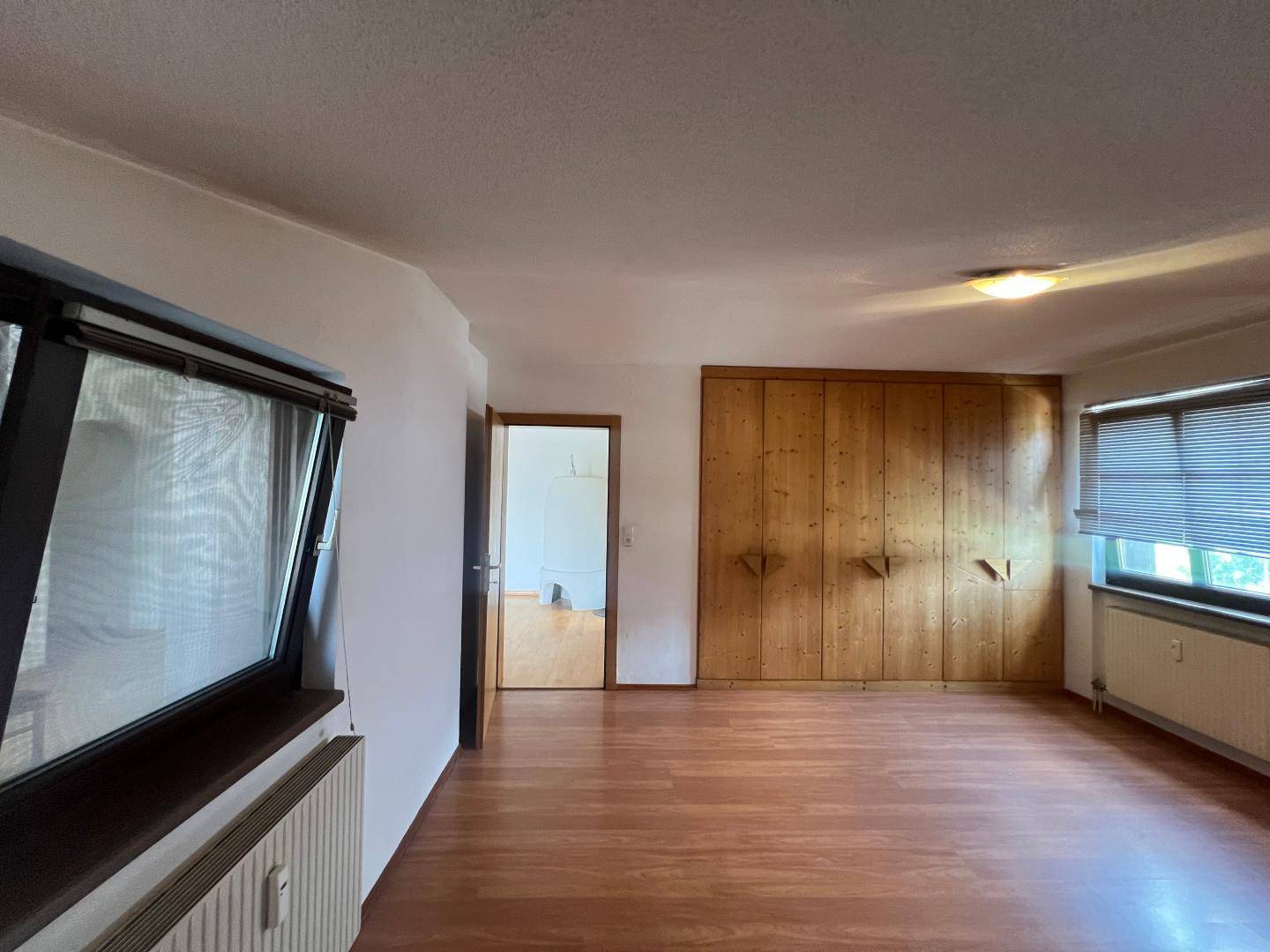 Geräumiges Schlafzimmer mit Einbauschränken aus Holz, Holzboden und Fenstern mit Jalousien.