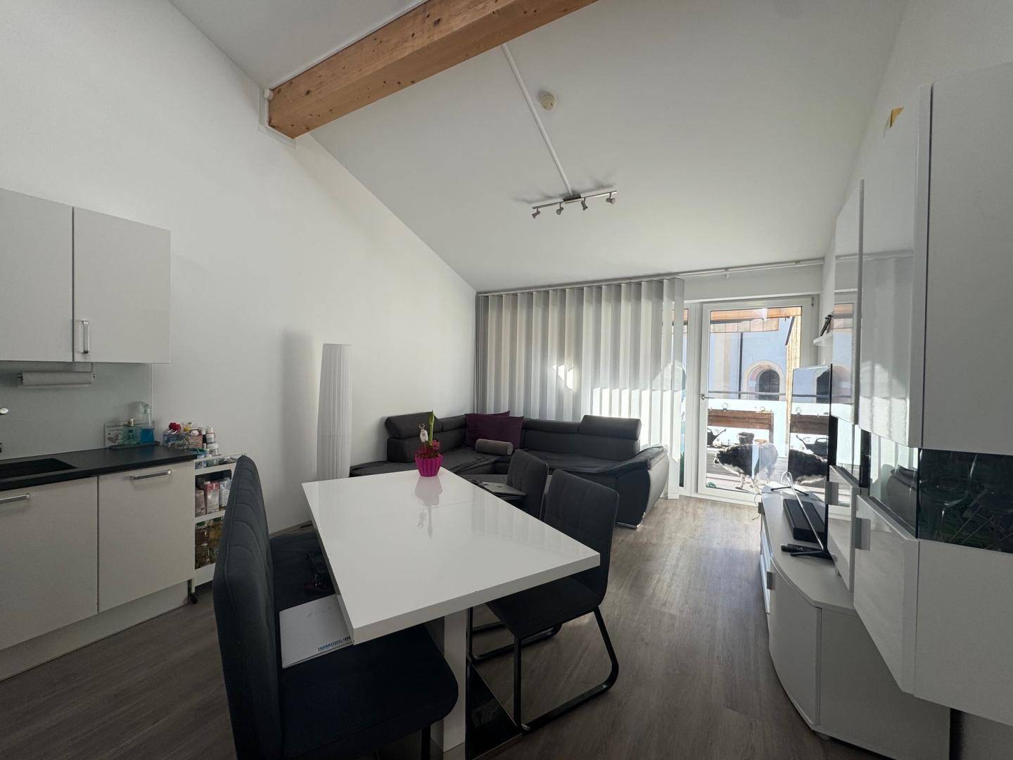 Heller offener Wohn-Essbereich mit integrierter Küche, Holzbalken und Zugang zum Balkon.