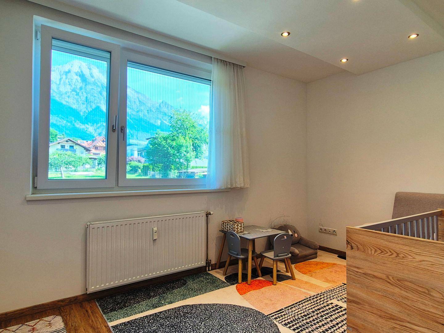 Kinderzimmer mit Babybett, kleinem Tisch und Stühlen, sowie einem Fenster mit Bergblick.