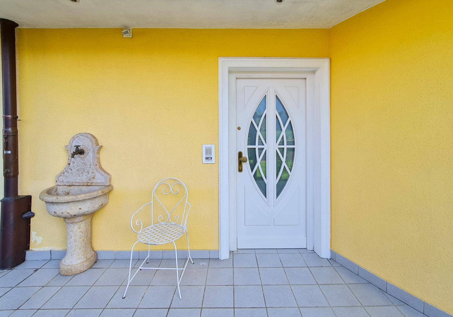 Eingangsbereich mit gelber Fassade, weißer Tür und einem dekorativen Steinbrunnen.