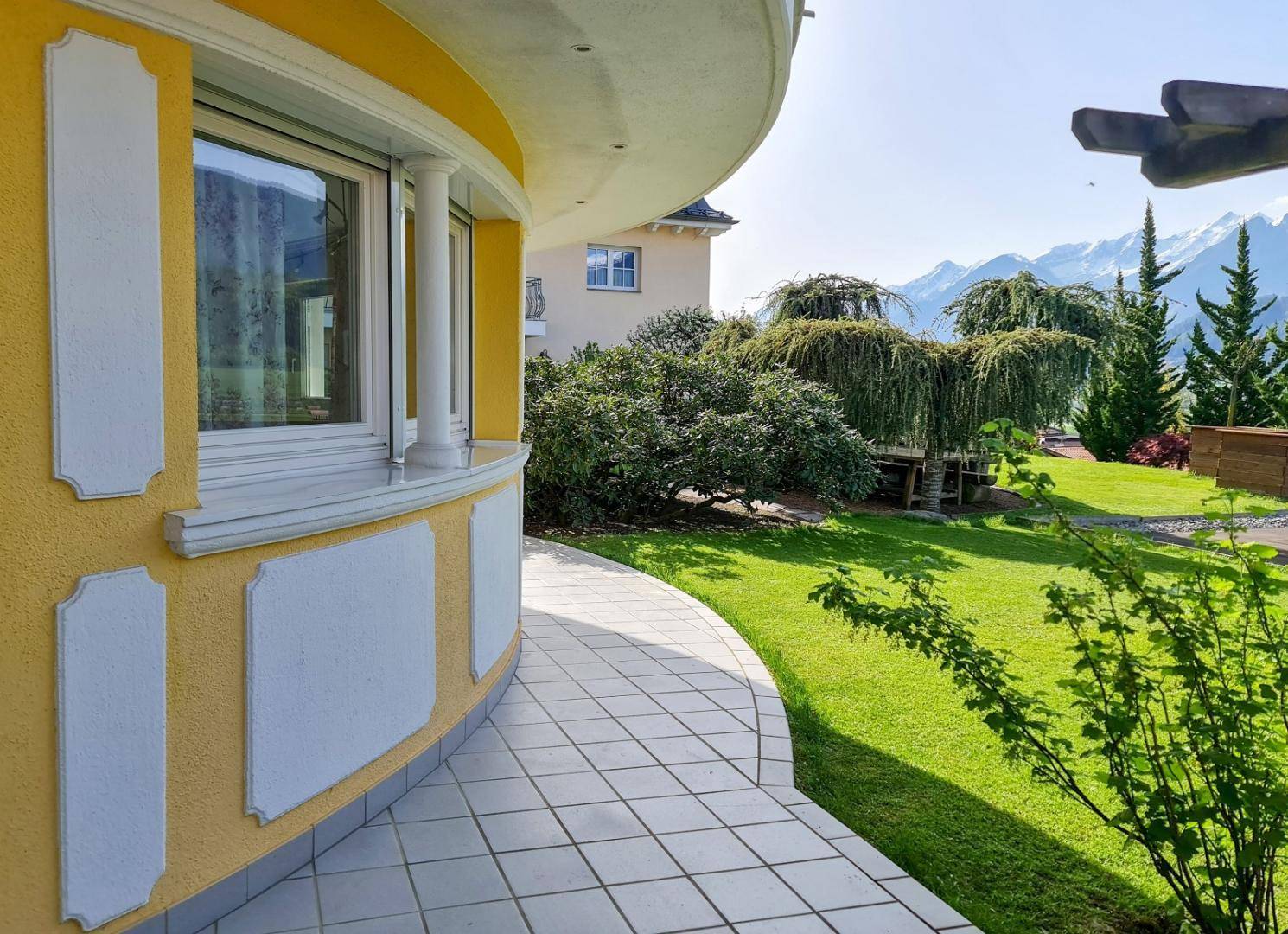 Gebogener Gebäudeteil mit großen Fenstern, umgeben von einem grünen Garten und Bergblick.