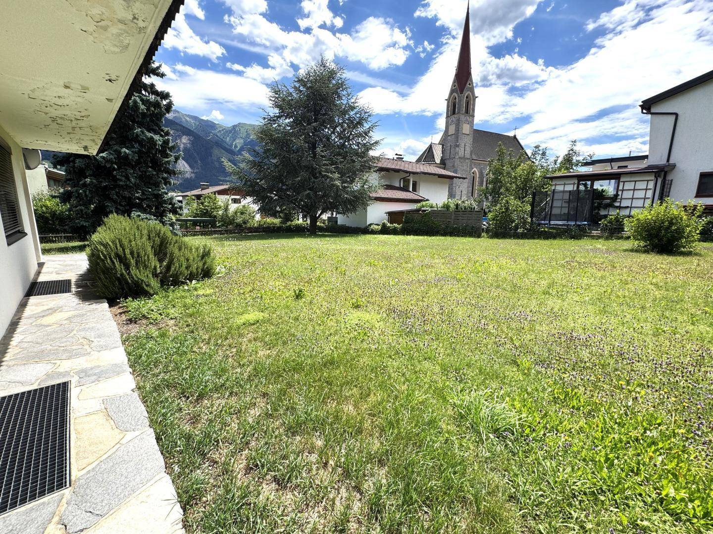Weitläufiger Garten mit Rasenfläche und Blick auf die nahegelegene Kirche und Berge.