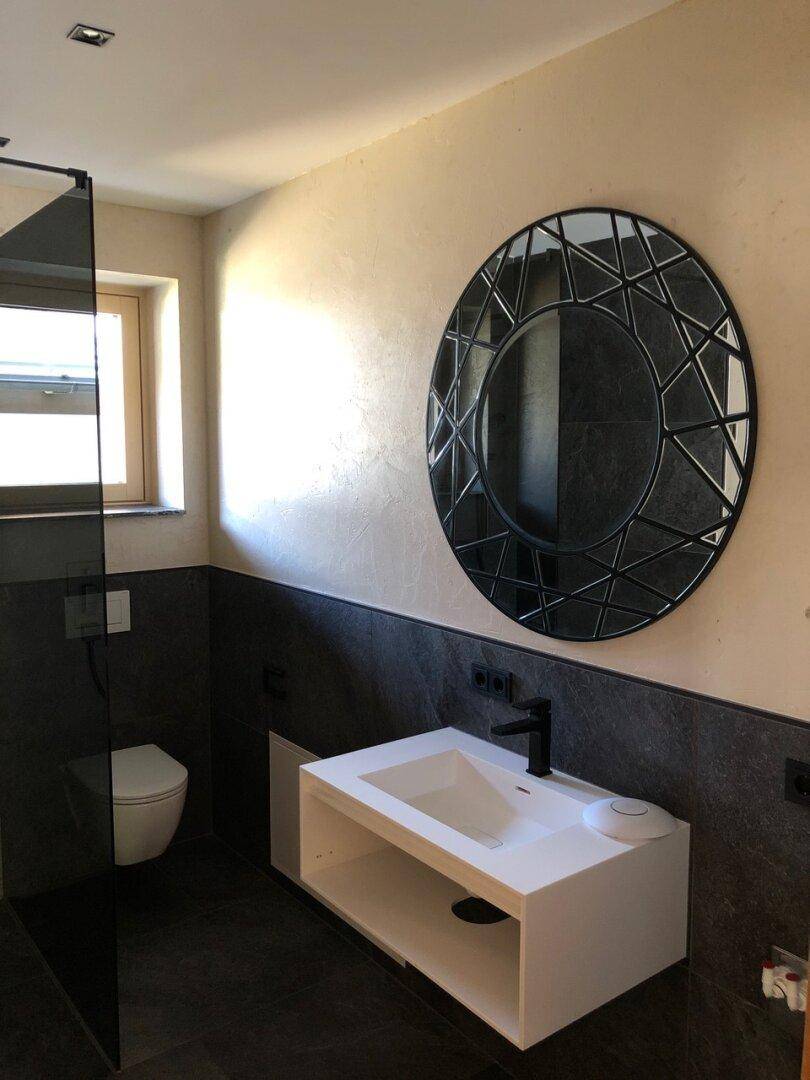 Modernes Badezimmer mit dunklen Fliesen, schwebendem Waschtisch, Wand-WC und einem dekorativen runden Spiegel.