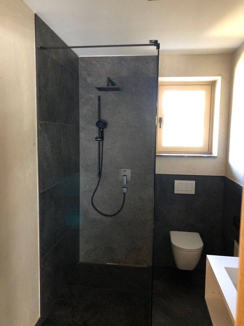 Modernes Badezimmer mit begehbarer Dusche, Wand-WC und dunklen Fliesen, die ein elegantes Ambiente schaffen.