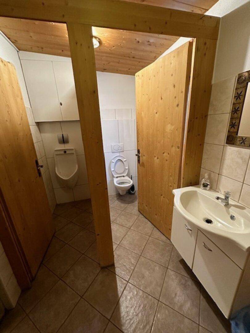 Saubere Gästetoilette mit Urinal, WC und Waschbecken, ausgestattet mit Fliesenboden und Holztüren.