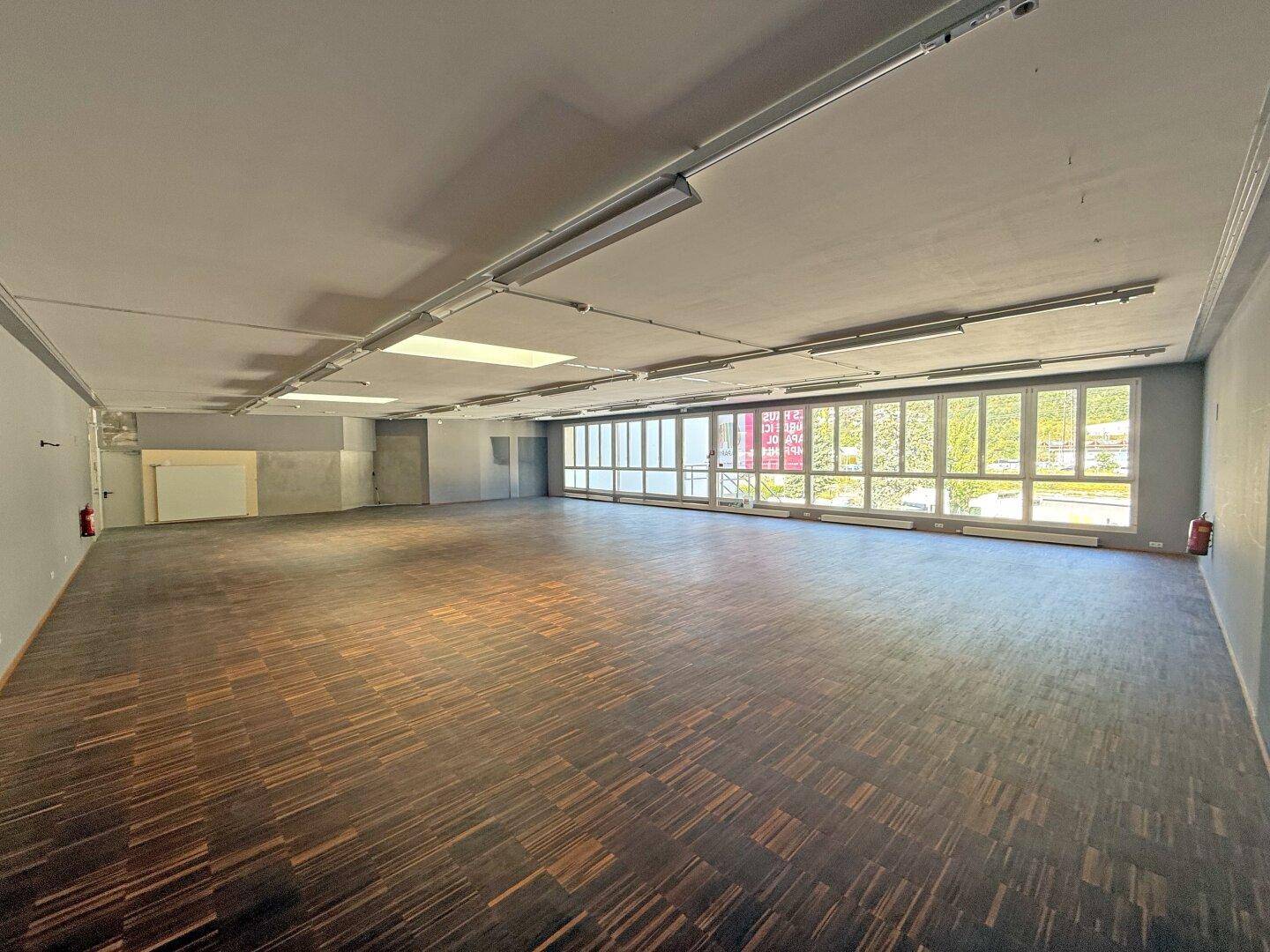 Großer, leerer Gewerberaum mit dunklem Holzparkett und breiter Fensterfront.