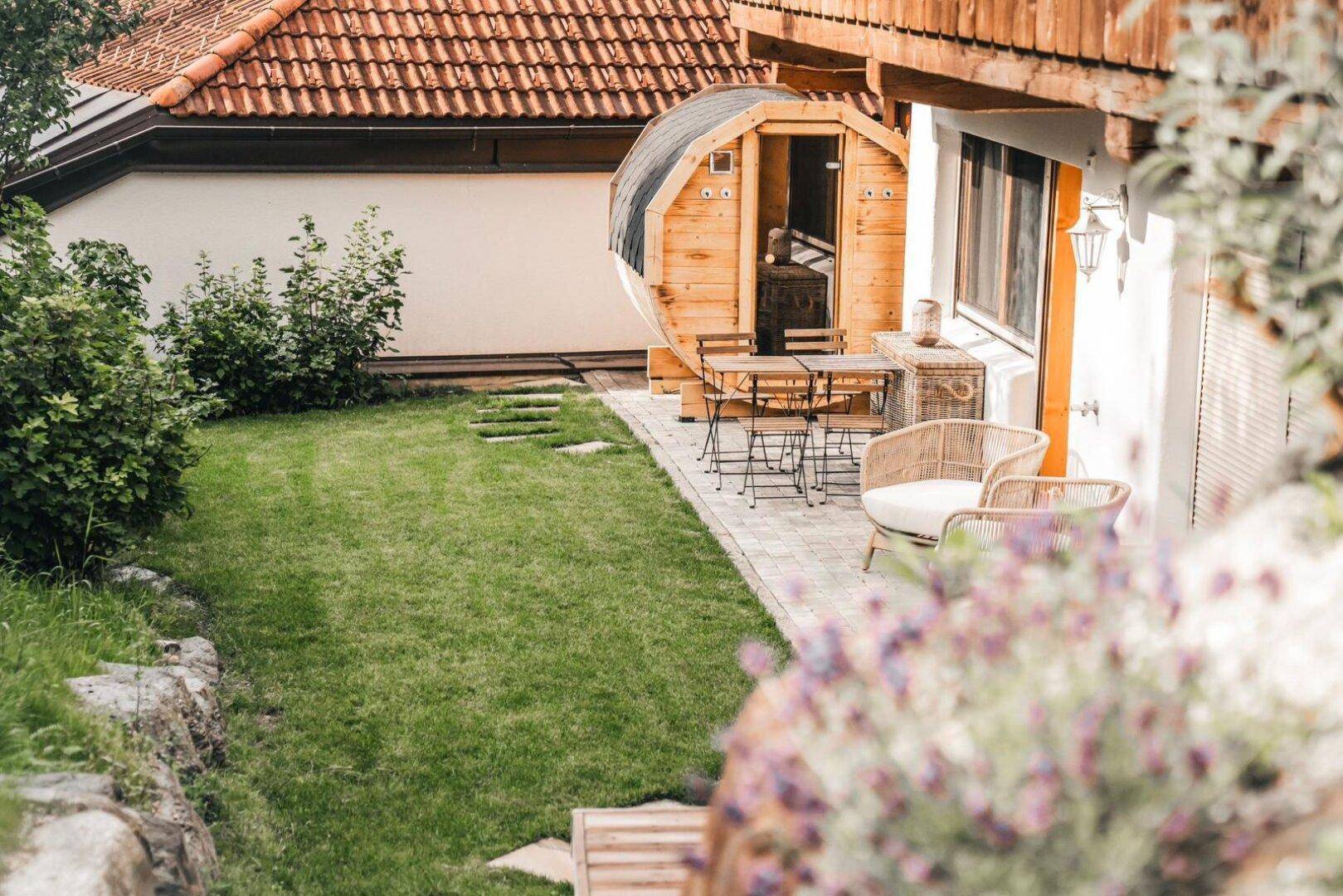 Kleiner Gartenbereich mit Fasssauna, gepflasterter Terrasse und Sitzgelegenheiten.