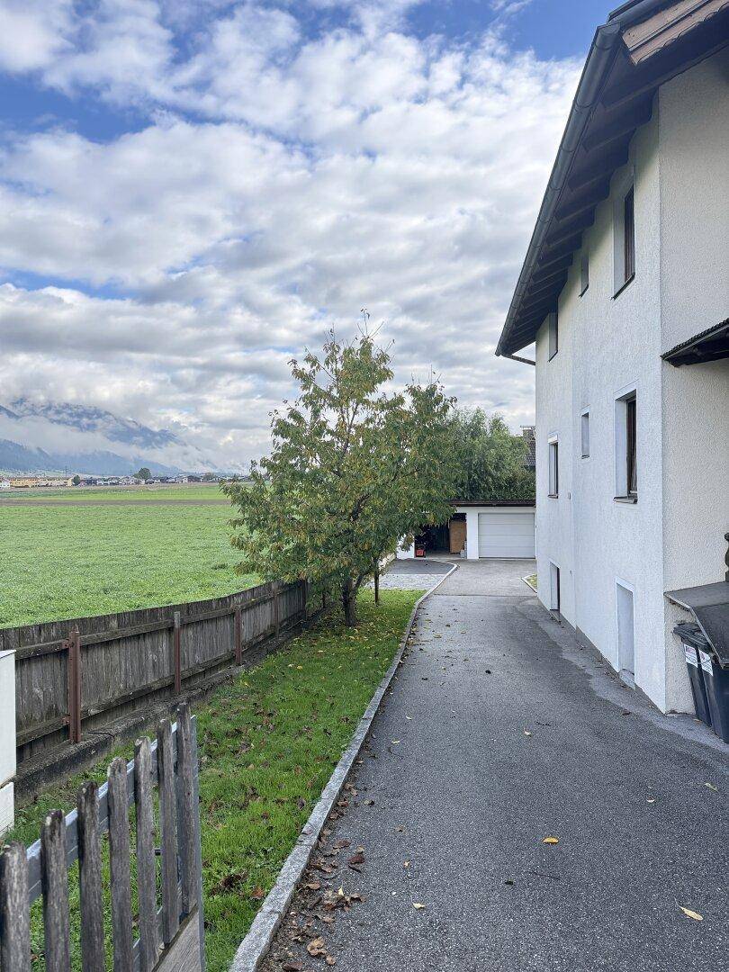Seitenansicht des Hauses mit Zufahrt, grüner Wiese und Bergen im Hintergrund unter bewölktem Himmel.