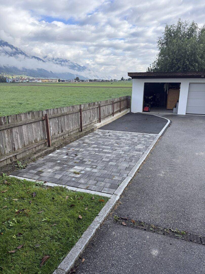 Gepflasterter Parkplatz und Garagenzufahrt neben einer grünen Wiese mit Bergblick.