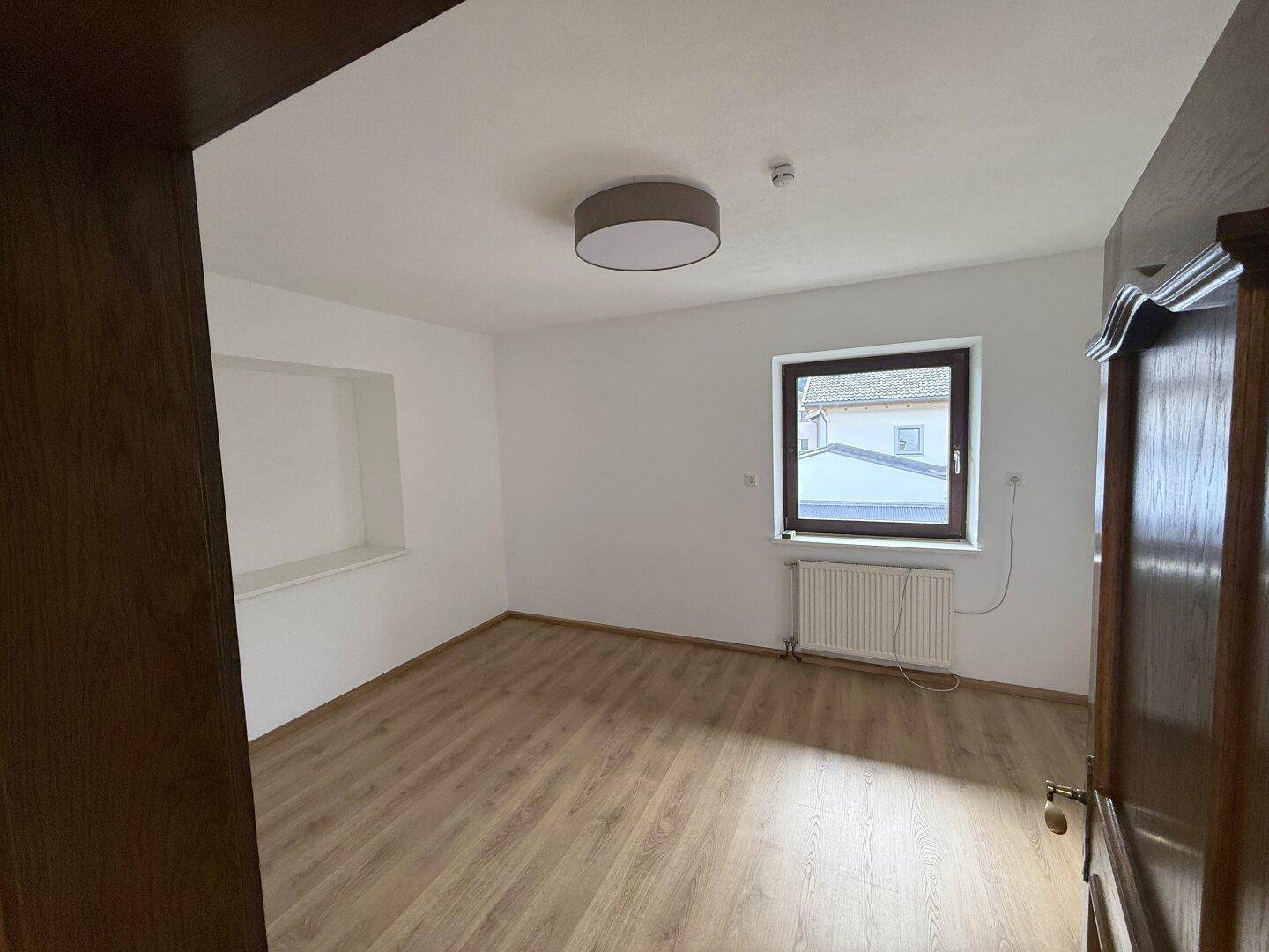 Schlafzimmer mit Laminatboden, weißen Wänden und einem Fenster mit Blick auf die Nachbarschaft.