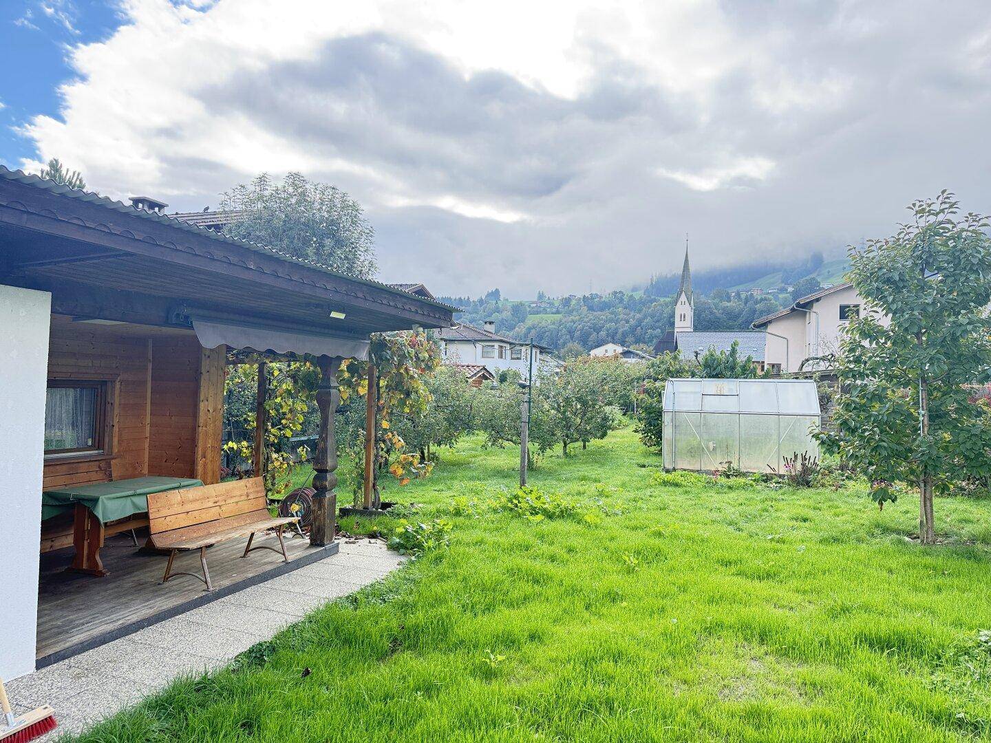 Grüner Garten mit überdachter Terrasse, Obstbäumen und Blick auf eine Kirche und Berge.