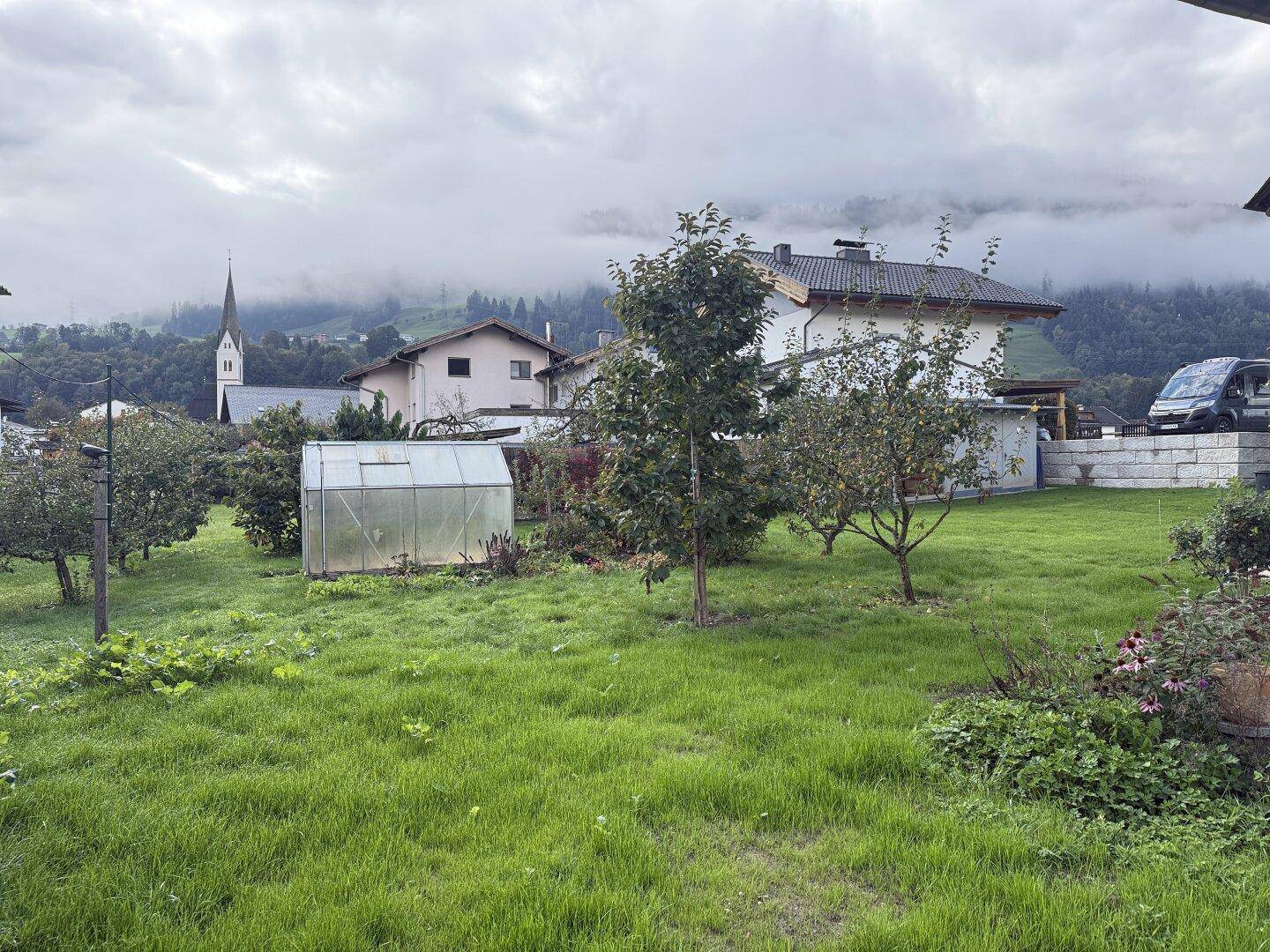 Weitläufiger grüner Garten mit Obstbäumen, Gewächshaus und Blick auf die umliegende Landschaft.