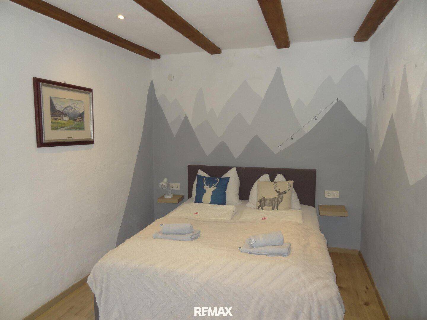 Gemütliches Schlafzimmer mit sichtbaren Holzbalken, einer modernen Bergwandmalerei und hellem Holzboden.