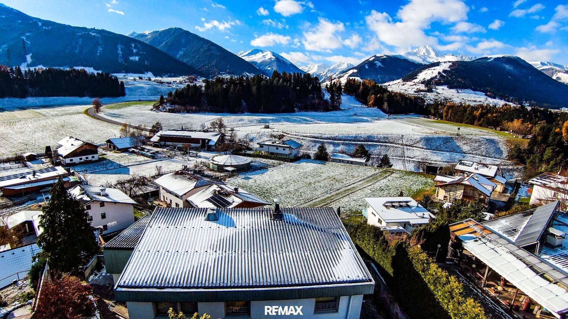 Luftaufnahme einer Wohnsiedlung in einer verschneiten Berglandschaft unter blauem Himmel.