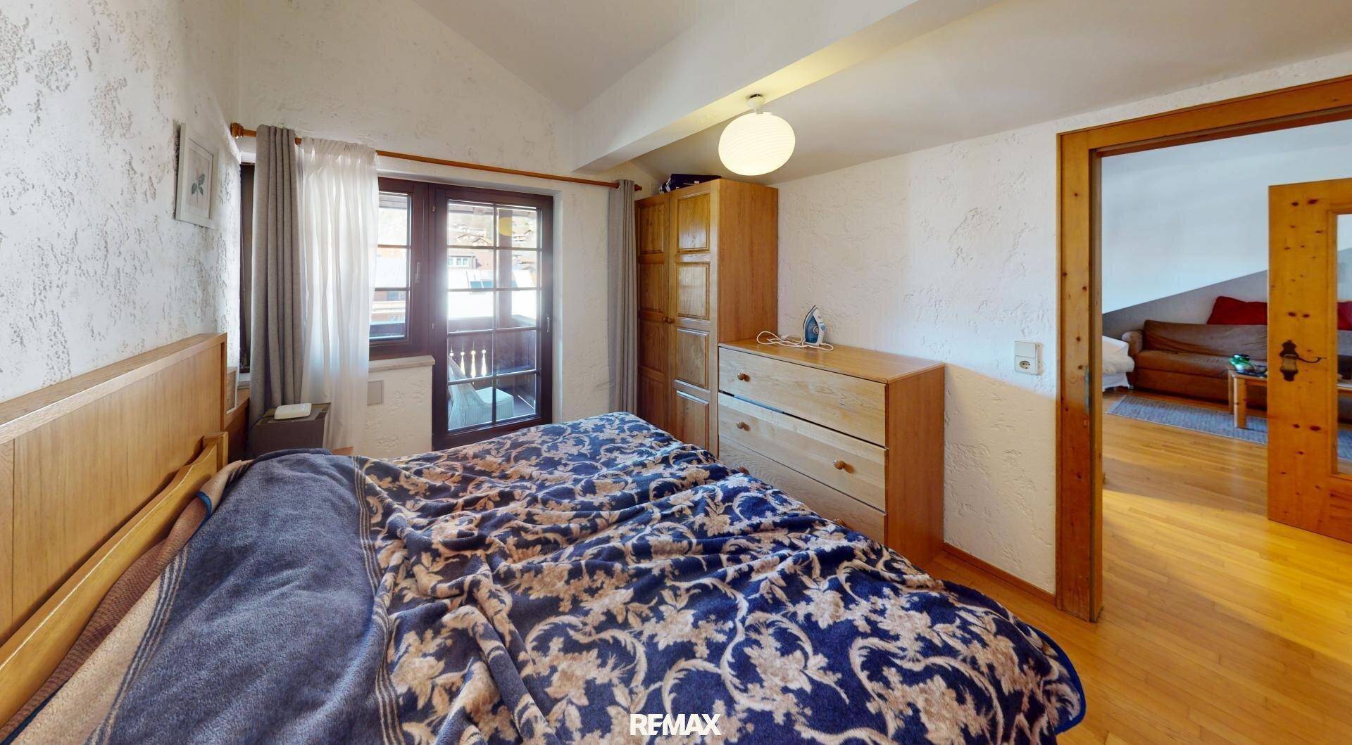 Schlafzimmer mit Doppelbett, Holzkommode und Zugang zu einem Balkon, mit Blick in einen weiteren Raum.