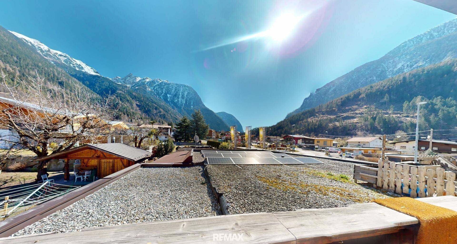 Panoramablick vom Dach mit Solaranlagen, umgeben von majestätischen Bergen und einem Dorf.