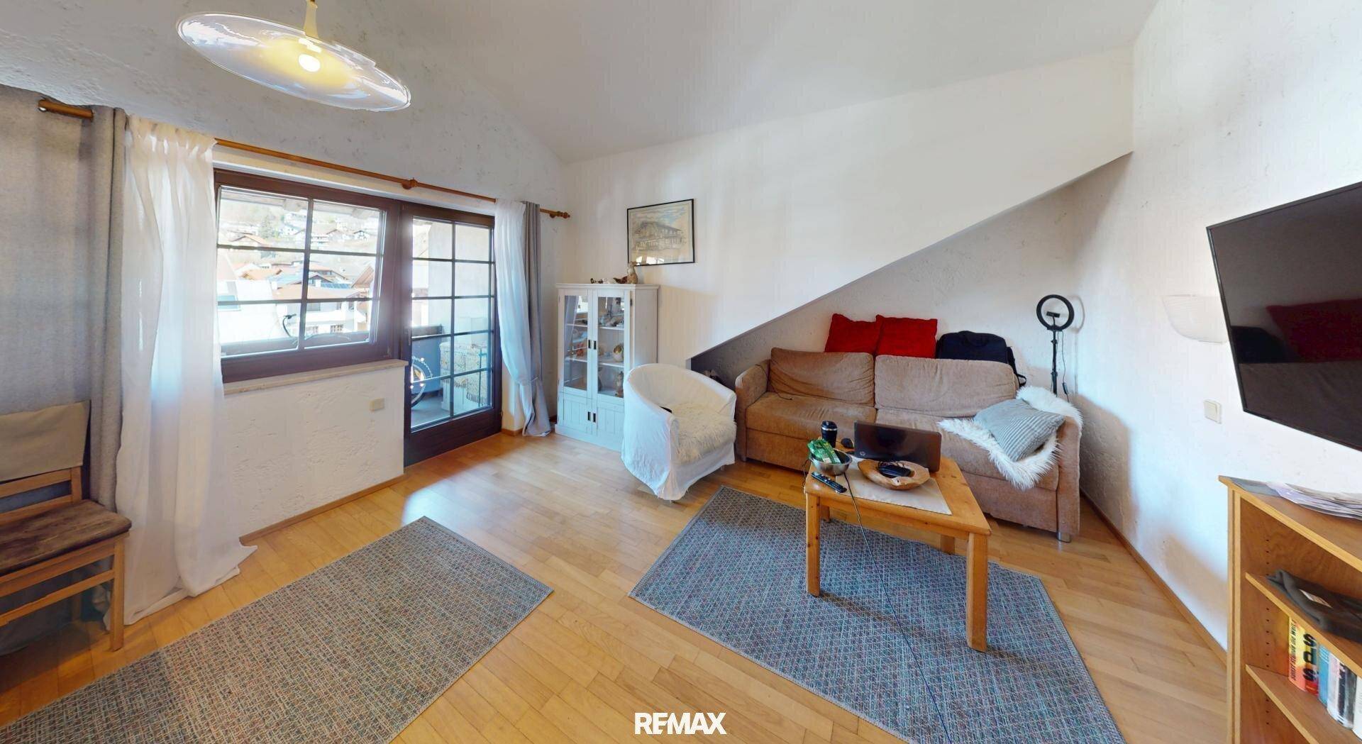 Helles Wohnzimmer mit Sofa, Fernseher und Zugang zum Balkon, unter einem Dachschrägenbereich.