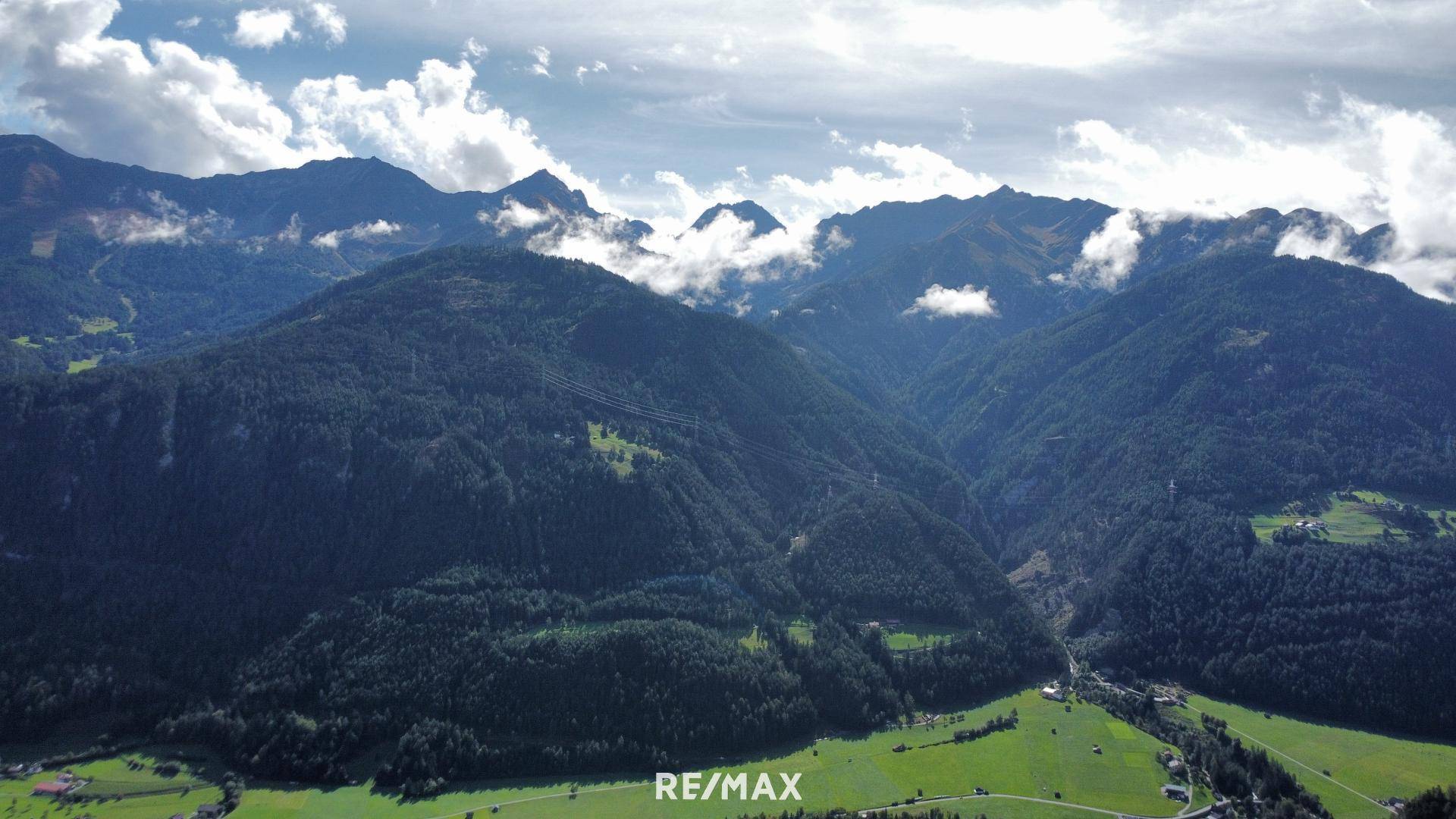 Weitläufige Ansicht von bewaldeten Bergen und einem Tal mit grünen Wiesen und kleinen Siedlungen.