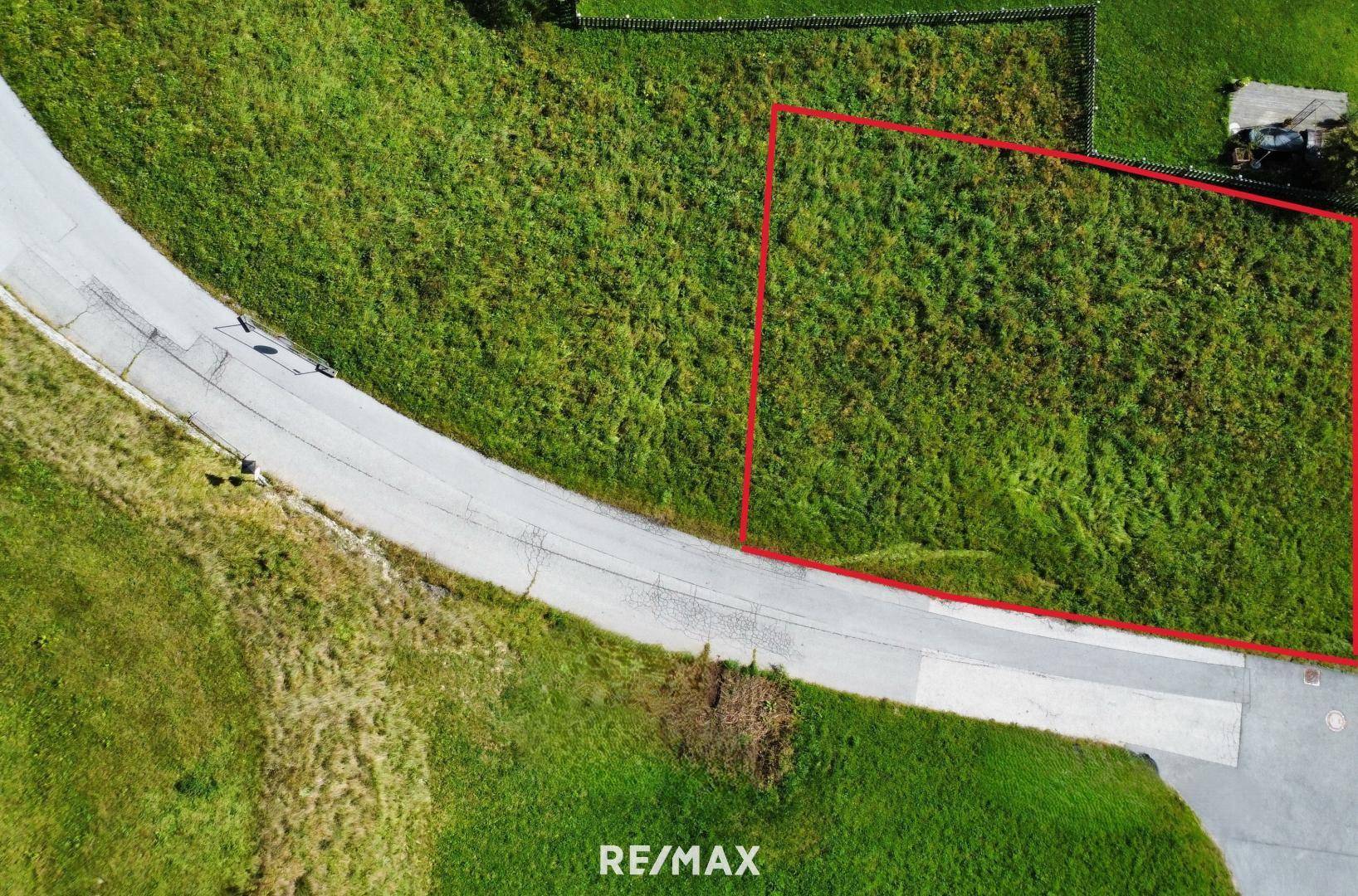 Luftaufnahme eines grünen Grundstücks, rot umrandet, neben einer Zufahrtsstraße in ländlicher Umgebung.