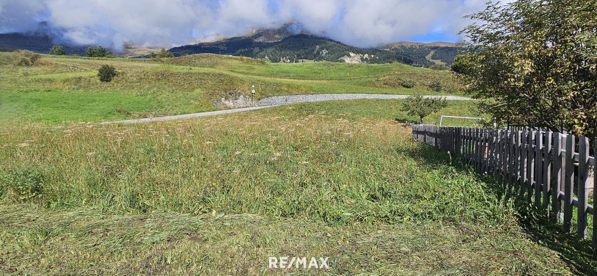 Weitläufige grüne Wiese mit einem Holzzaun und einer Straße, umgeben von sanften Hügeln und Bergen.