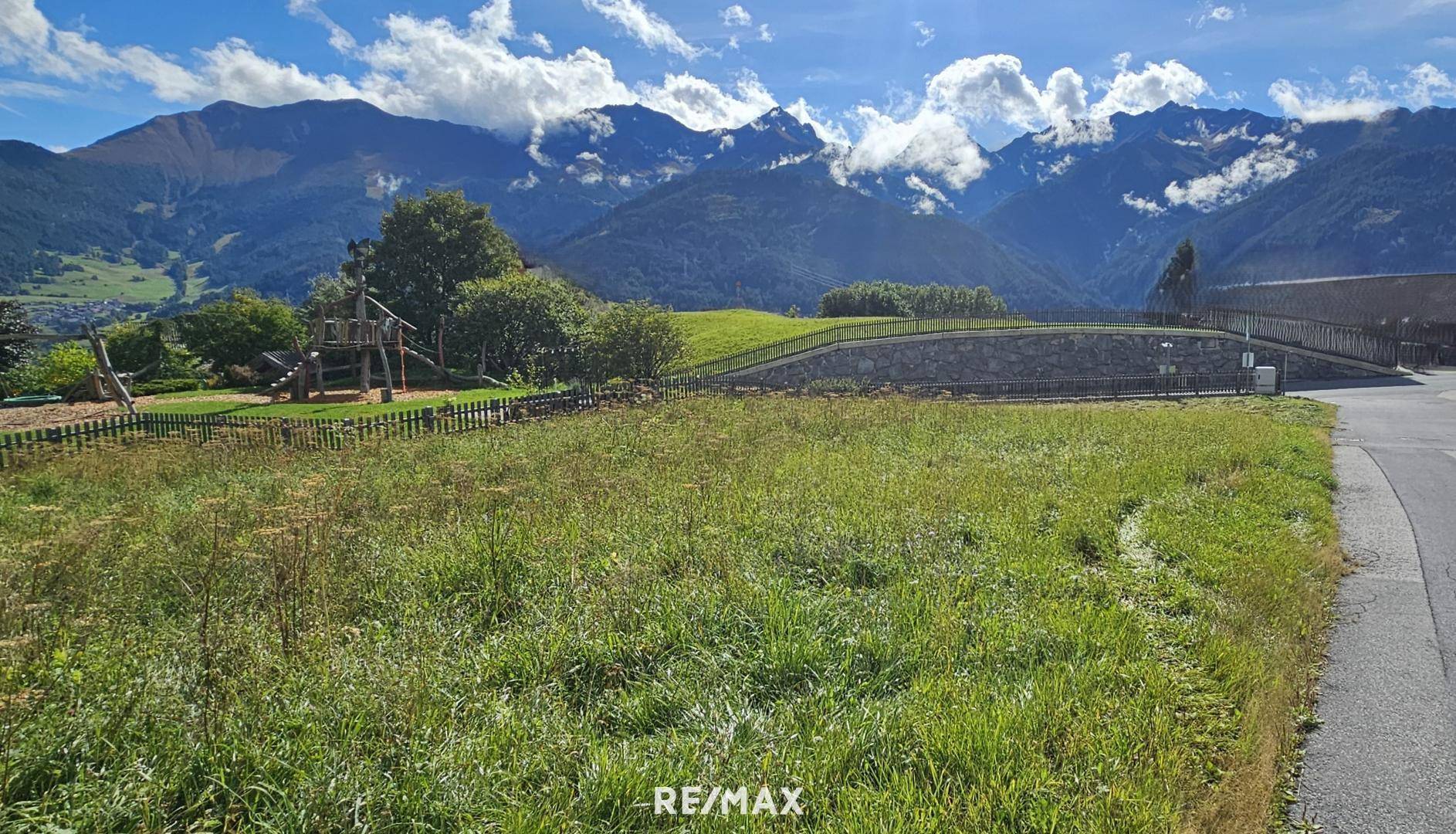 Grüne Wiese mit Spielplatz im Vordergrund und majestätischen Bergen im Hintergrund unter blauem Himmel.