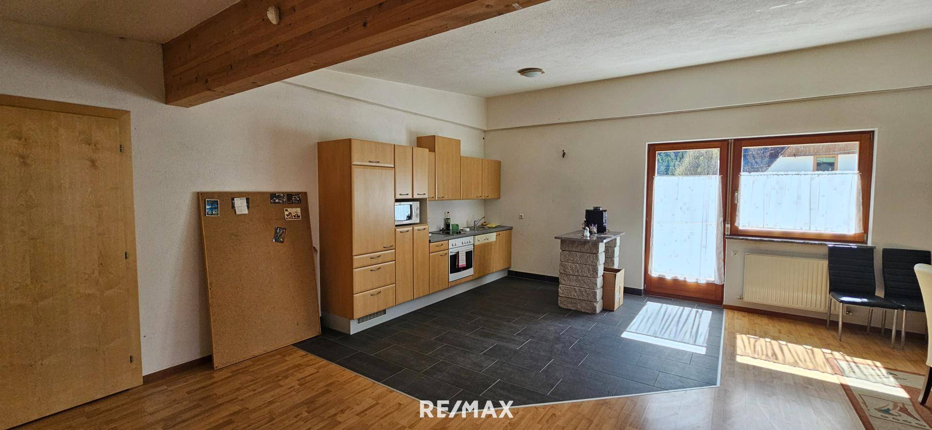 Offener Wohnbereich mit Holzküche, dunklen Fliesen und Holzboden, ergänzt durch sichtbare Deckenbalken und große Fenster.