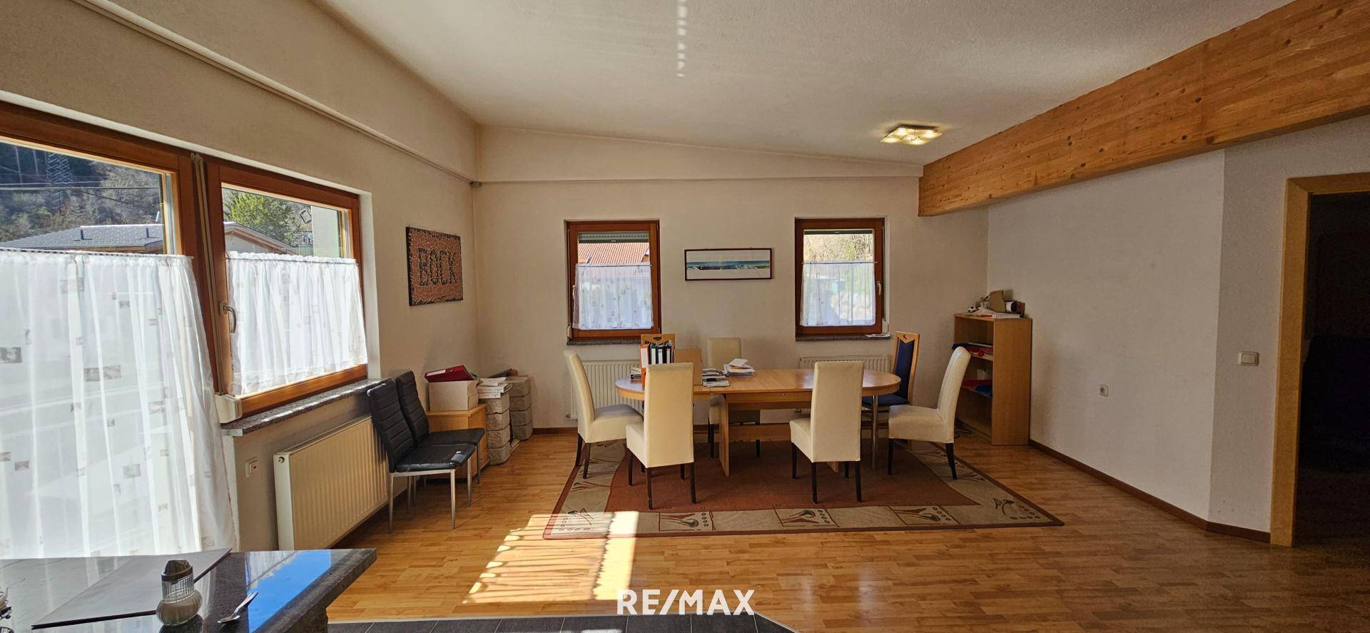 Heller Essbereich mit Holztisch und Stühlen, großen Fenstern und sichtbaren Deckenbalken, angrenzend an einen Wohnbereich.