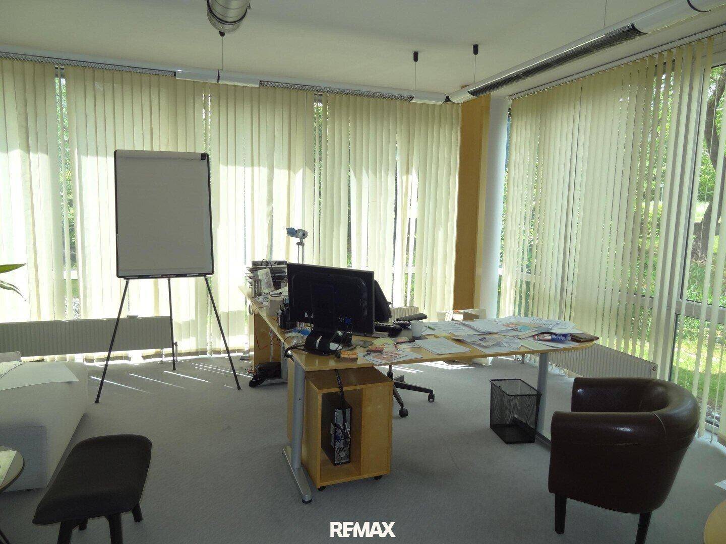 Helles Büro mit großem Schreibtisch, Flipchart und vertikalen Jalousien an den bodentiefen Fenstern.