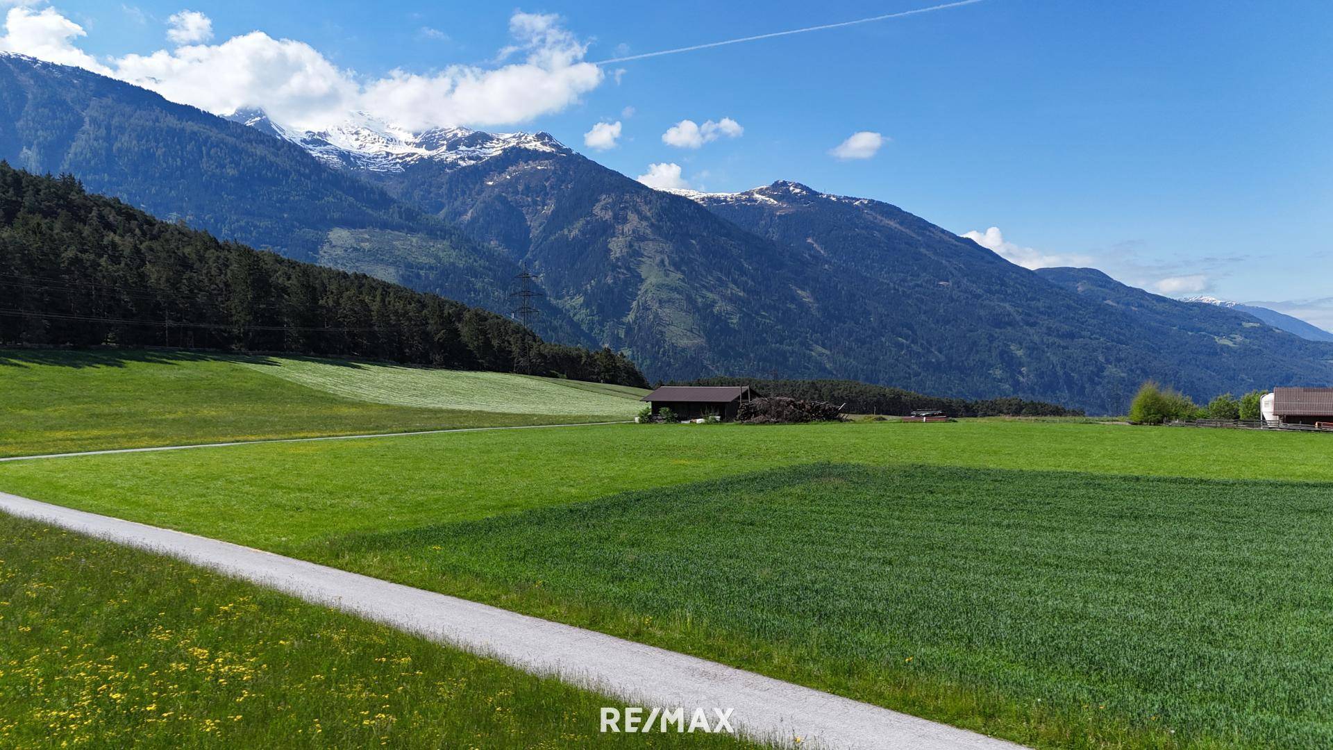 Weitläufige grüne Felder mit einem Bauernhaus im Hintergrund und majestätischen Bergen unter blauem Himmel.