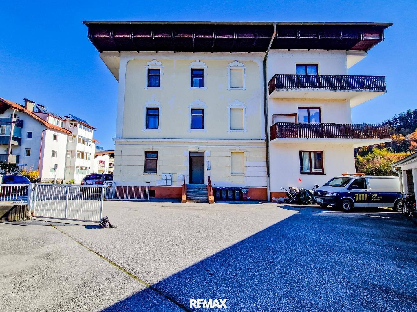 Älteres Mehrfamilienhaus mit Balkonen und Parkplätzen, umgeben von Bergen.