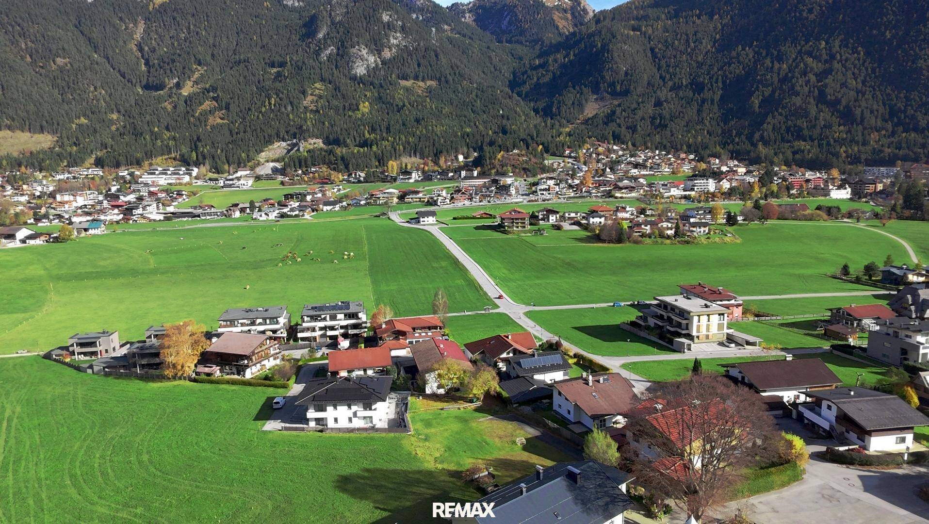 Ausgedehnte Luftaufnahme einer Siedlung in einer grünen Talschaft mit bewaldeten Bergen im Hintergrund.