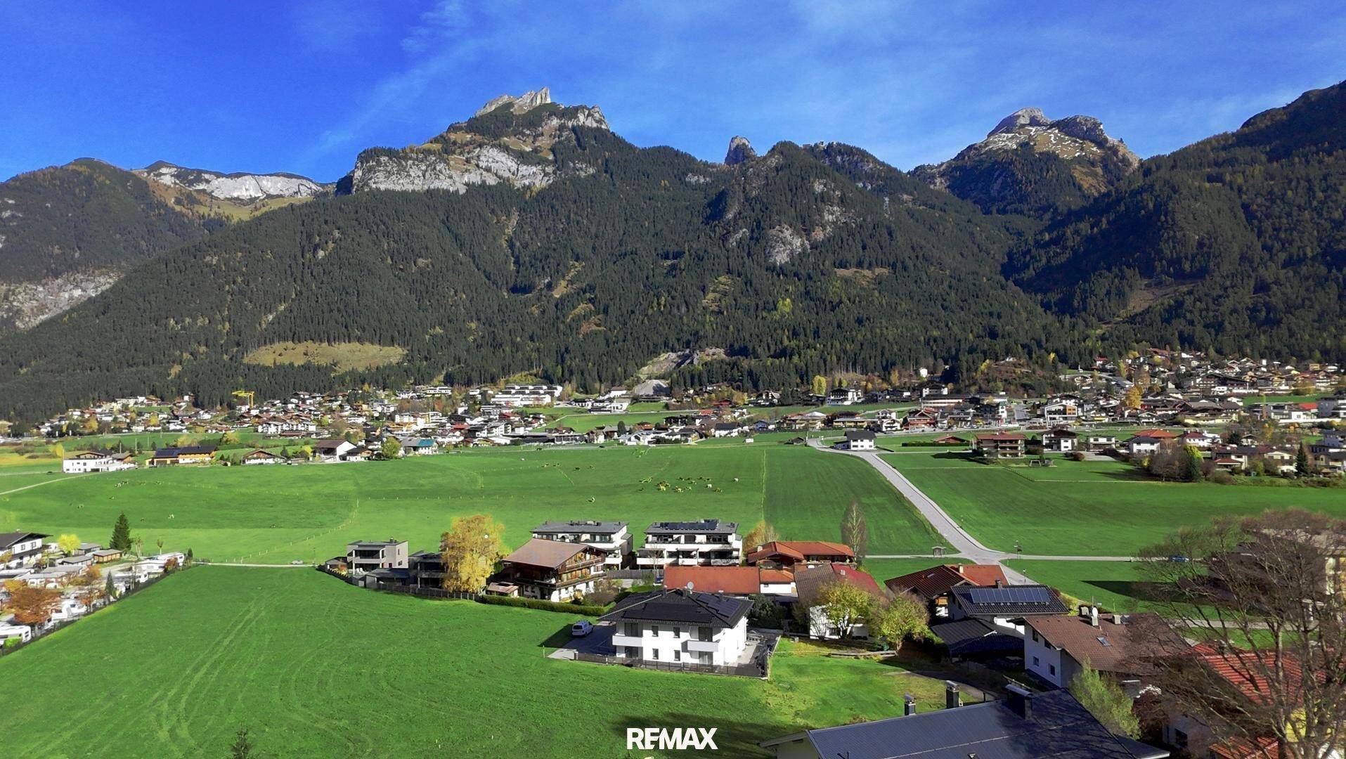 Weitläufige Luftaufnahme eines Dorfes in einem Tal, umgeben von grünen Wiesen und majestätischen Bergen.