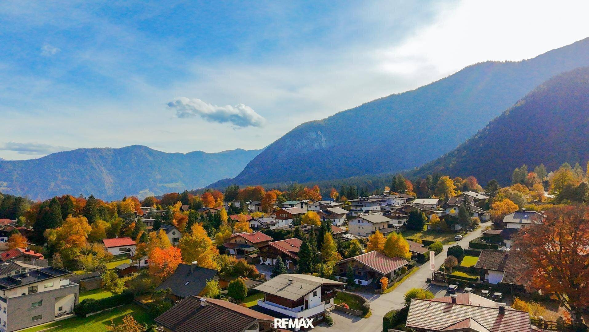 Malerische Herbstlandschaft eines Dorfes mit bunten Bäumen und Bergen unter strahlend blauem Himmel.
