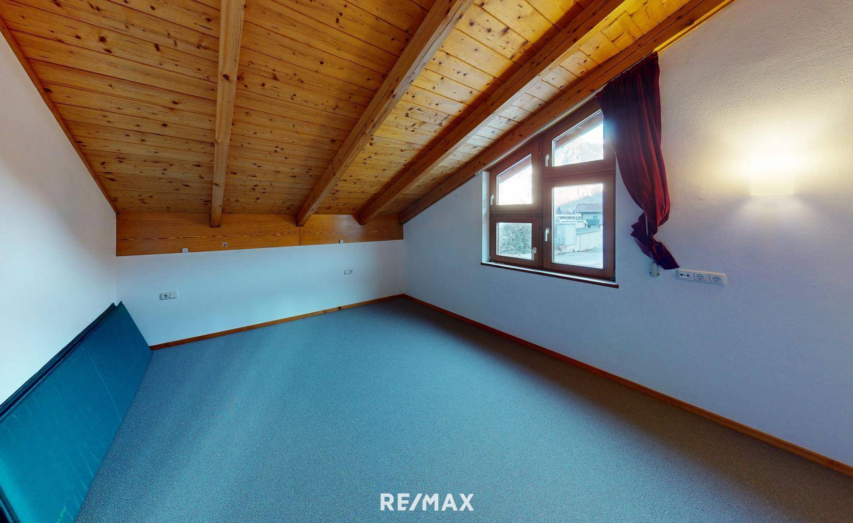 Dachzimmer mit Holzbalkendecke, Teppichboden und Fenster mit Blick nach draußen.