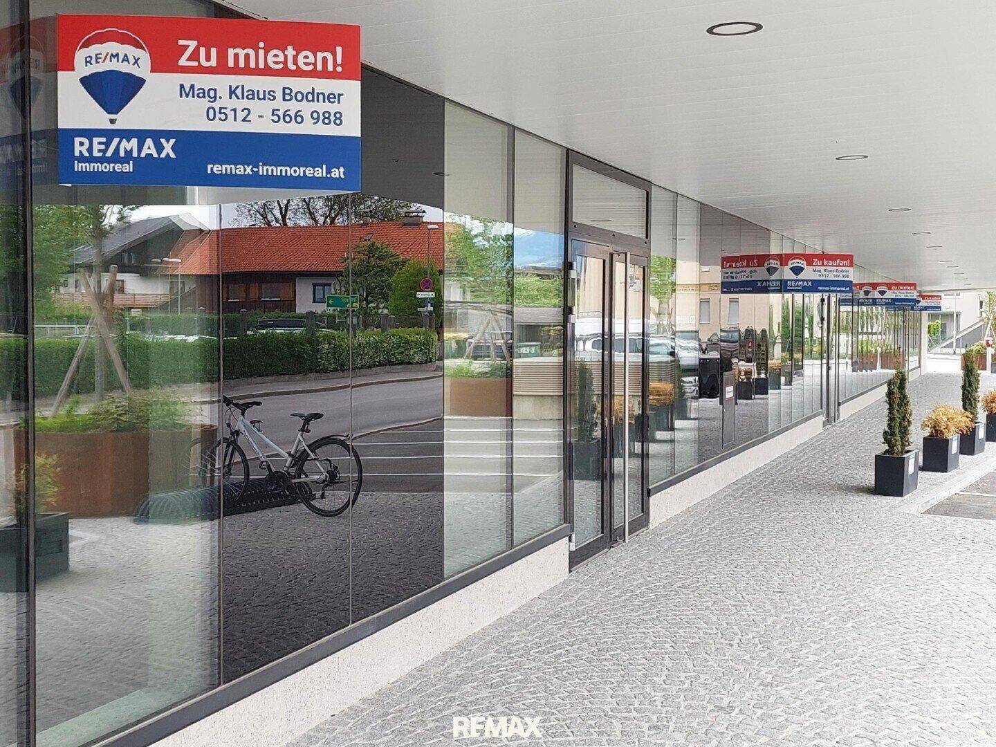 Moderne Glasfassade eines Geschäftsgebäudes mit reflektierenden Fenstern, gepflastertem Gehweg und Vermietungsschildern.